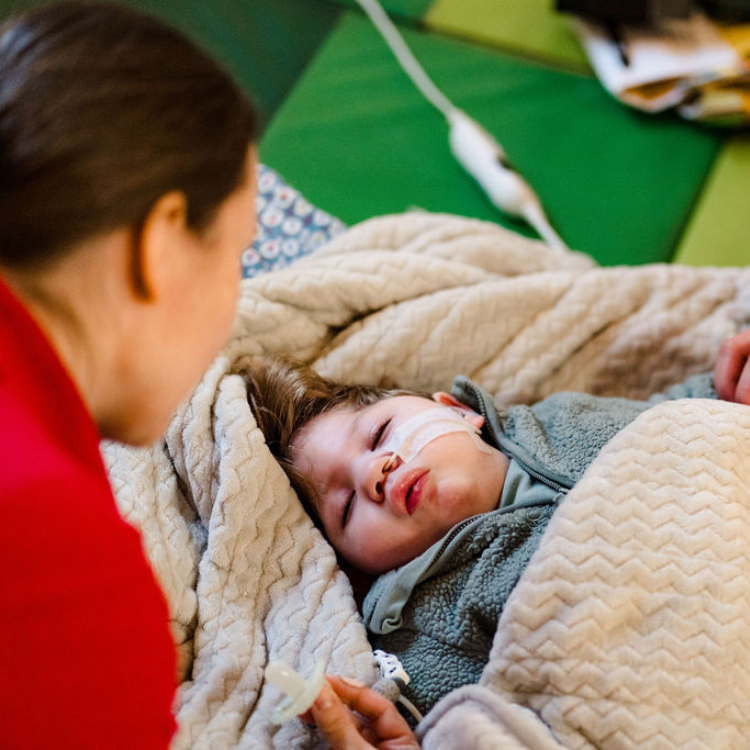 Young boy with a feeding tube laying down, with a lady holding his hand and looking at him.